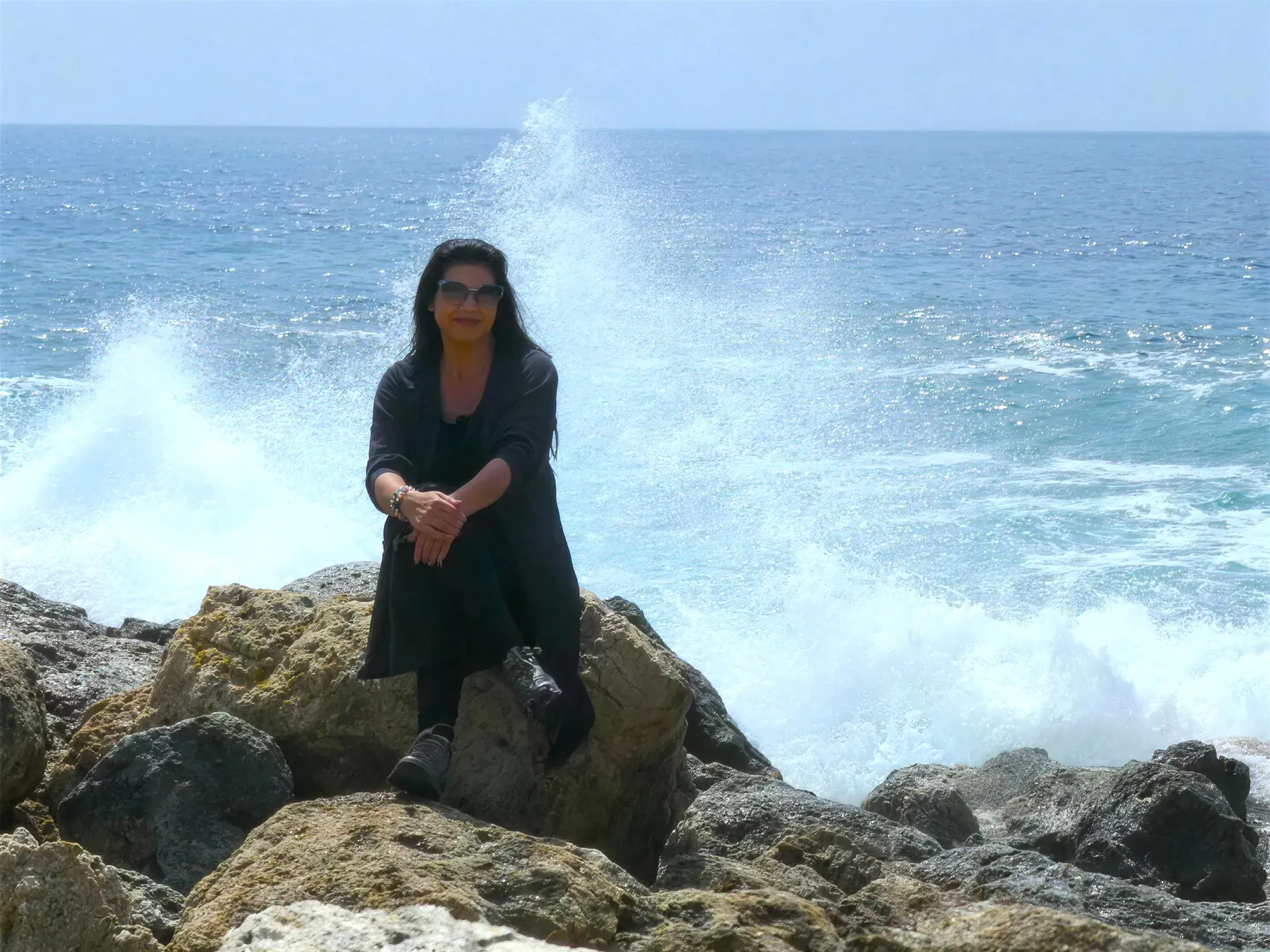 Sabine Kuhn sitzt gelassen auf Felsen am Meer, während hinter ihr eine mächtige Welle aufbricht