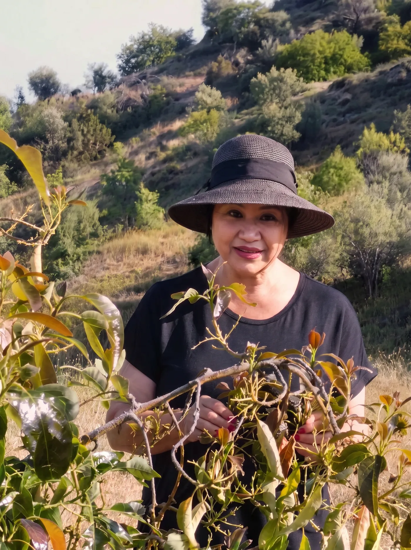 Sabine Kuhn mit Sonnenhut lächelt freundlich zwischen Avocadozweigen in der zyprischen Hügellandschaft