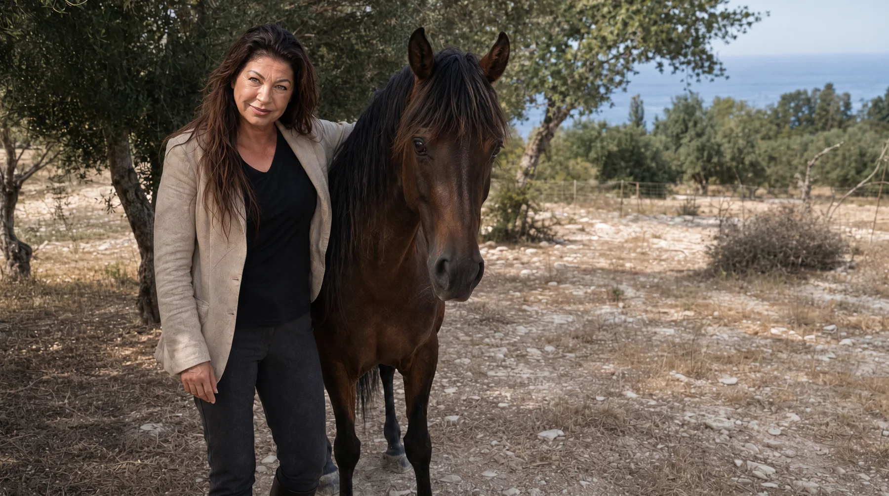Sabine Kuhn steht mit einem ihrer Pferde im Olivenhain, im Hintergrund glitzert das Mittelmeer
